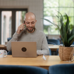 Ein Mann mit Kopfh&ouml;rer sitzt an einem Holztisch vor einem Laptop und l&auml;chelt w&auml;hrend eines Videoanrufs.